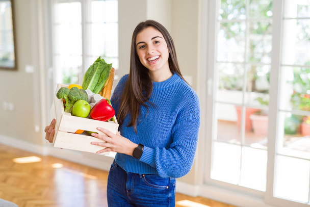 Молодая женщина улыбается держа деревянную коробку с продуктами из доставки
 - Фото, изображение