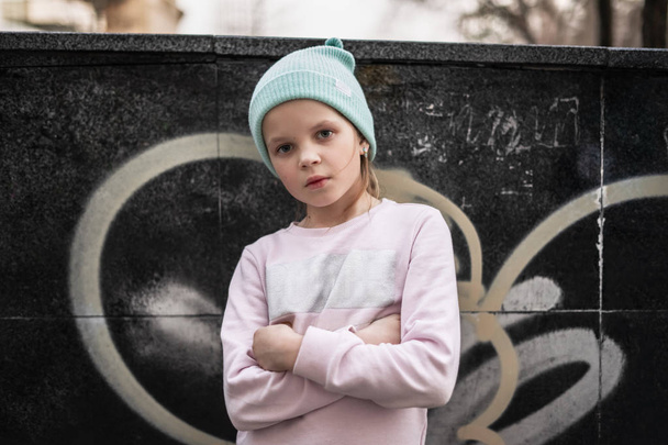 A serious child girl folded her arms across her chest against the background of a city wall with graffiti elements. Street portrait of a real person. - Fotografie, Obrázek