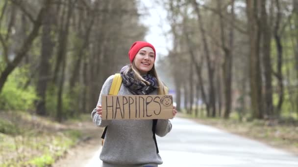 Uśmiechnięta Autostopem dziewczyna na drodze z plakatem szczęścia. Życie w podróży - Materiał filmowy, wideo