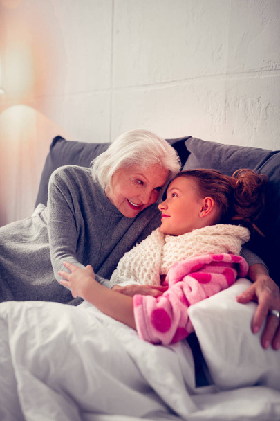 Caring granny hugging her cute girl staying in bed feeling sick - Photo, image