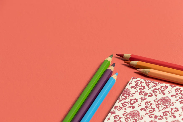 fondo coral vivo color del 2019 con cuaderno con patron renacentista en rojo burgund y beige en una esquina y acompaado de lpices de colores naranja, rosa, rojo, azul, can y verde - Foto, Bild