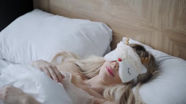 Young woman sleep on comfortable bed in a mask for sleeping. Blindfold on eye. Morning in hotel room. White pillow and blanket - Footage, Video