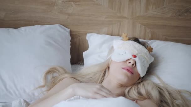 Mladá žena spí na pohodlné posteli v masce na spaní. Zavázané oči. Dobré ráno v hotelovém pokoji. Bílý polštář a přikrývka - Záběry, video