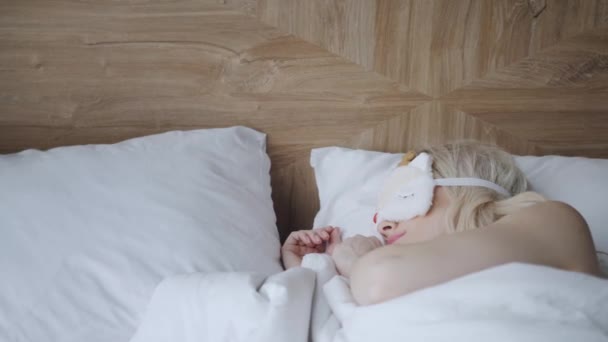 Young woman sleep on comfortable bed in a mask for sleeping. Blindfold on eye. Morning in hotel room. White pillow and blanket - Filmagem, Vídeo