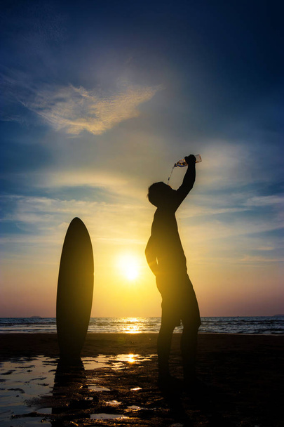 Sörf adam silueti şişeden su içmek bir sörf tahtası ile standı. Gün batımı sahilinde sörf yapıyorum. Açık su sporları macera yaşam tarzı. Yaz aktivitesi. 20'li yıllarda Asya erkek modeli - Fotoğraf, Görsel