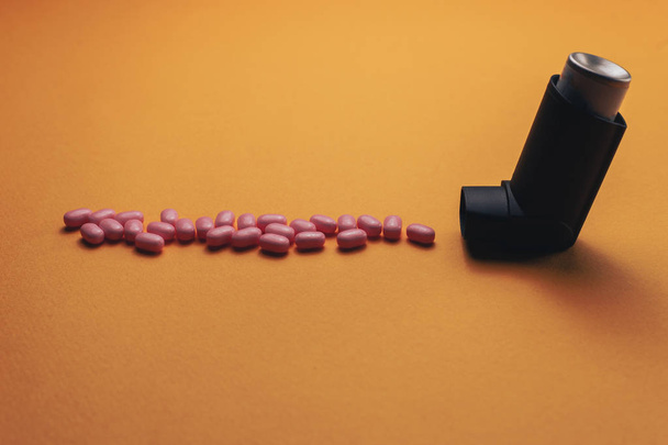 schwarzer Asthma-Inhalator mit Aerosolpartikeln aus rosa Pillen (Vitamine, Farbstoffe, Geschmacksverstärker, Nahrungsergänzungsmittel, Bonbons) auf orangefarbenem Hintergrund.  - Foto, Bild