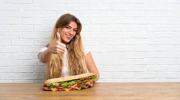 jong blond vrouw holding een Groot sandwich met duim omhoog - Foto, afbeelding