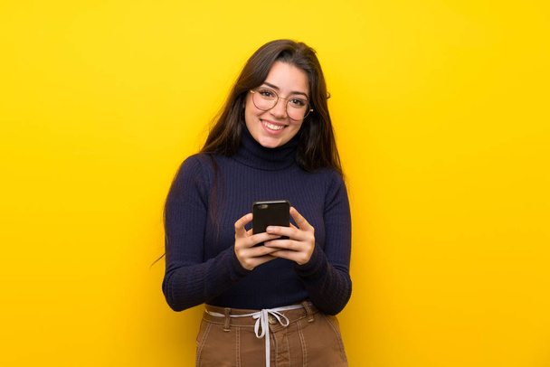 Dívka z puberty přes izolovanou žlutou zeď, která posílá zprávu s mobilním - Fotografie, Obrázek