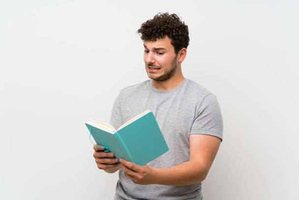 Άνθρωπος με σγουρά μαλλιά πάνω από απομονωμένο τοίχο κρατώντας και διαβάζοντας ένα βιβλίο - Φωτογραφία, εικόνα
