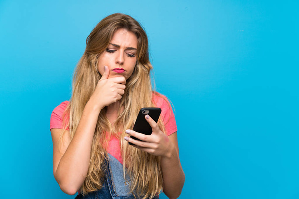 Nuori blondi nainen haalarit yli eristetty sininen seinä ajattelu ja lähettää viestin
 - Valokuva, kuva