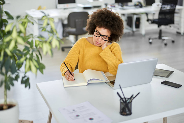 Attractive mixed race businesswoman dressed casual taking notes in notebook while sitting in modern office. On desk laptop, tablet and smart phone. - Foto, Bild