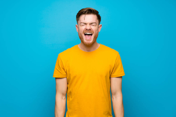 Redhead man over blauwe muur schreeuwen naar de voorkant met mond wijd open - Foto, afbeelding