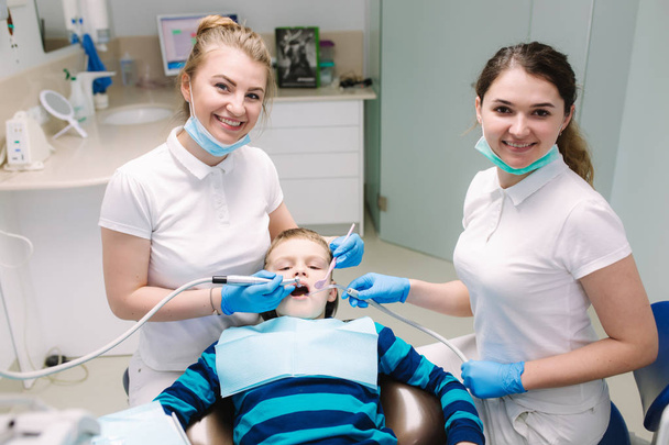 Dzieci dentysta i asystent egzaminu chłopcy zęby, a on siedzi na krześle dentystycznym w biurze. Kobieta dentysta - Zdjęcie, obraz