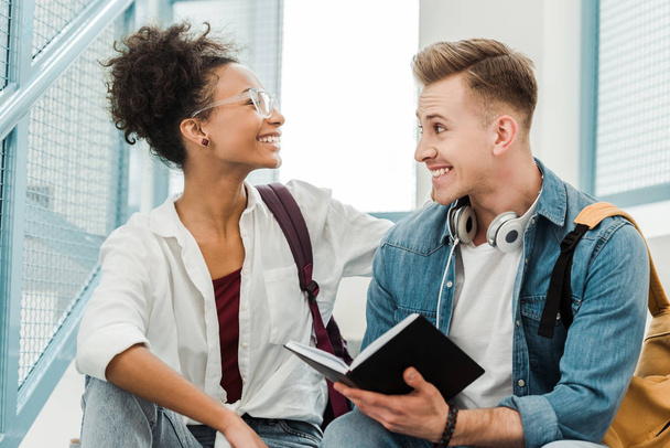 dos estudiantes multietnicos con cuaderno sonriendo en la universidad
 - Foto, Imagen