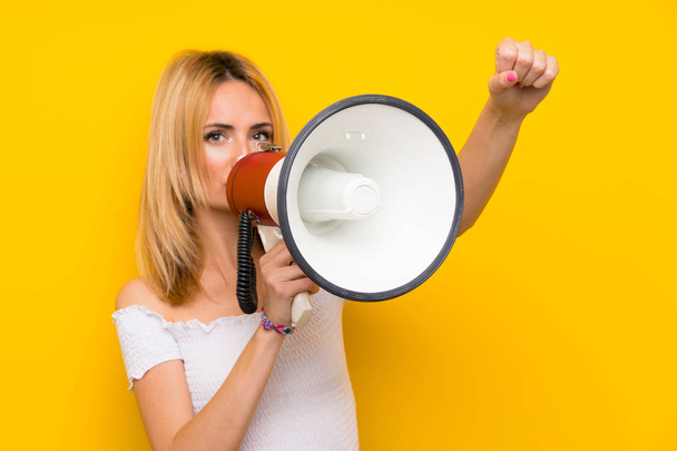 jonge blonde vrouw over geïsoleerde gele muur schreeuwen door een megafoon - Foto, afbeelding