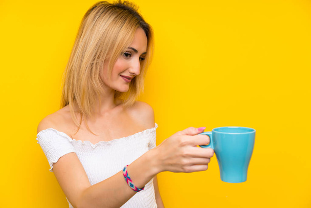 Młoda blondynka nad odizolowaną żółtą ścianą trzyma gorącą filiżankę kawy - Zdjęcie, obraz