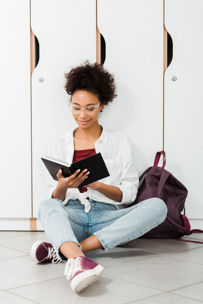 estudiante afroamericano sentado en el suelo y leyendo un cuaderno en el pasillo
 - Foto, Imagen