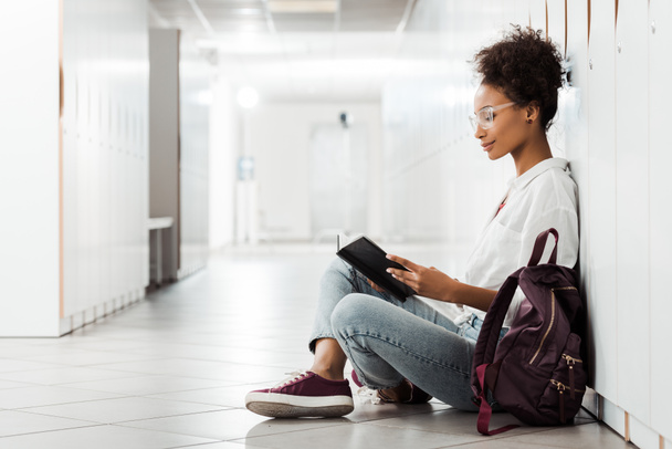 side view of african american student sitting on floor and reading notebook in corridor - Fotoğraf, Görsel