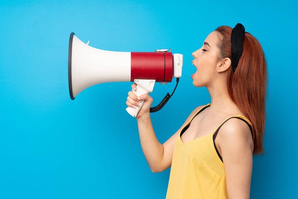 Young redhead woman over blue background shouting through a megaphone - Photo, Image