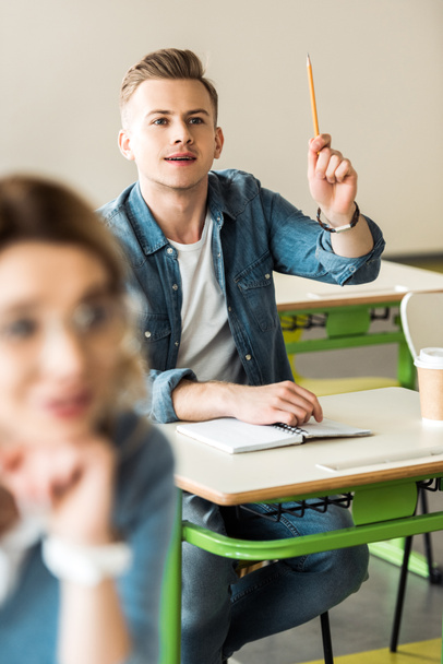 estudante de camisa jeans sentado na mesa e levantando a mão com lápis
 - Foto, Imagem