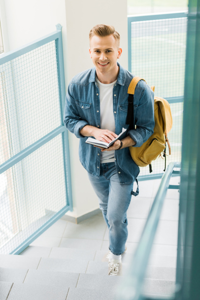 student in denim shirt with yellow backpack holding notebook in university - Zdjęcie, obraz