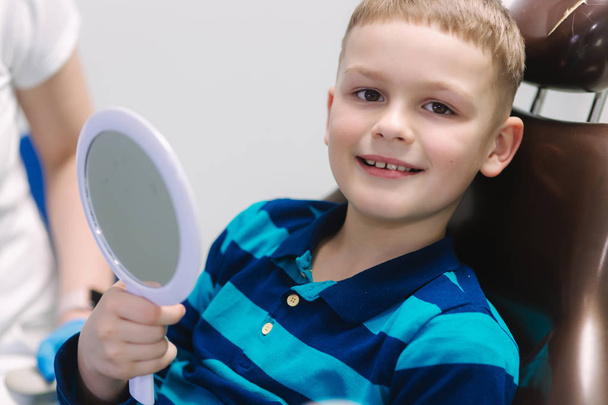 鏡を持ってダンティストの椅子に座っている小さな男の子。男性患者は彼の歯を見る - 写真・画像
