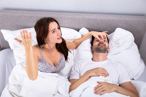 Häiriintynyt nuori nainen pitelee miehensä nenää estääkseen häntä kuorsaamasta sängyssä
 - Valokuva, kuva