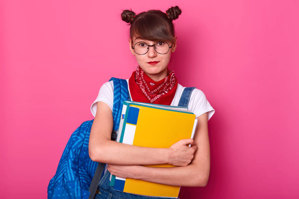 Student girl embraces colorful paper folder isolated on pink background, lady ready for serious work at university, wearing white t shirt, red bandana on neck and denim overalls. Education concept. - Foto, Imagem