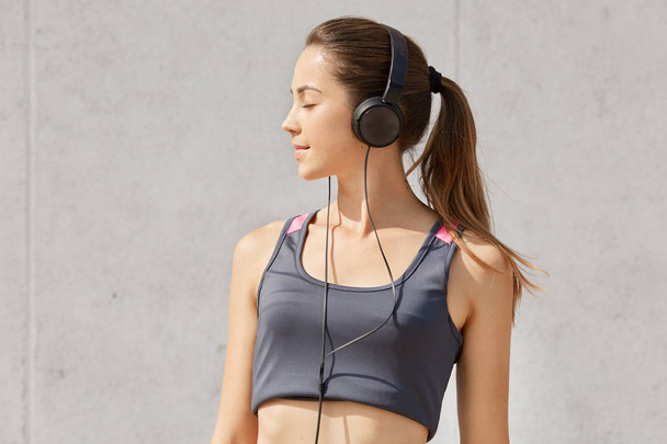 Portrait of caucasian pretty woman listening to music while having workout in gym, has headphones, posing in studio, stands with closed eyes, isolated over gray background. Health care concept. - Fotoğraf, Görsel