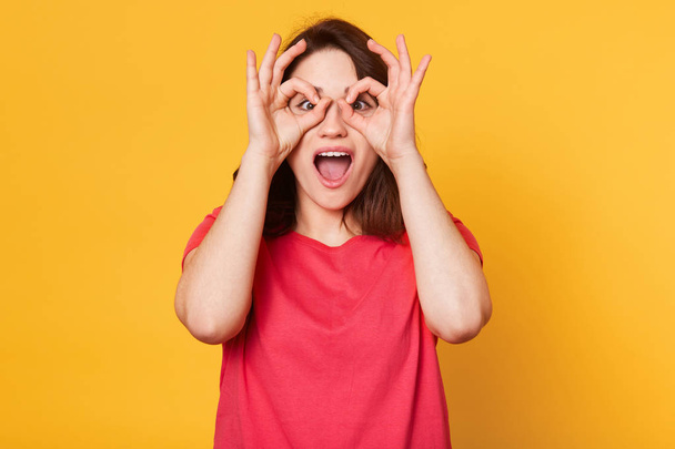 Image of young happy woman in red t shirt, holding her hand over her eyes as glasses and looking through fingers, having fun while being photographed in studio, isolated over yellow background. - Photo, Image
