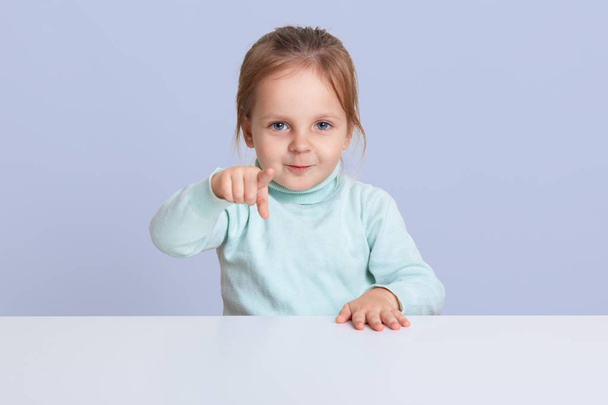 Half length studio photo of charming little girl wearing casually, pointing directly at camera, showing something to her mother, choosing doll, model posing isolated over blue studio background. - Foto, Imagem