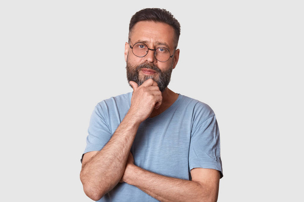 Portrait of middle bearded aged male with pensive facial expression, dressed gray cassual t shirt and round spectacles, keeps hand under chin, looks thoughtful, thinks abot new idea, has great plans. - Photo, image