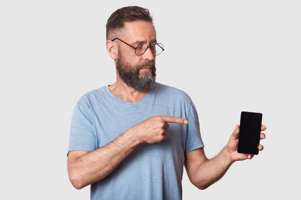 Close up portrait of attractive middle aged male in gray casual t shirt, stands against white wall, pointing with fore finger on blank screen of his smartphone, looking on device. Copy space. - Fotó, kép