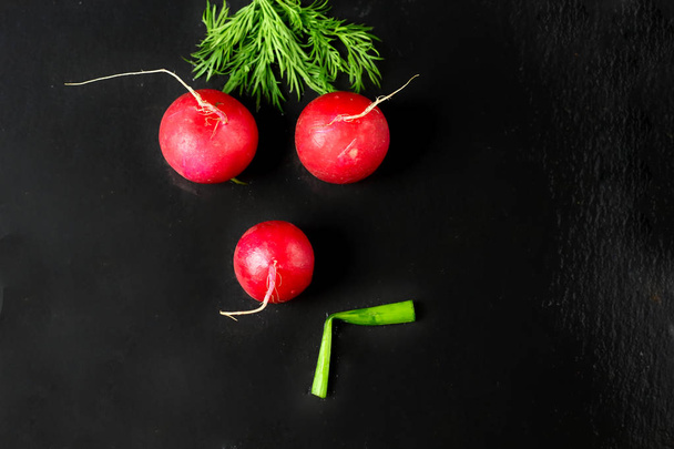 Abstract funny serious unhappy sad face with dill hair made of vegetables, red radish and green leek smiling on a black background. Healthy eating vegetarian organic nutrition - Zdjęcie, obraz