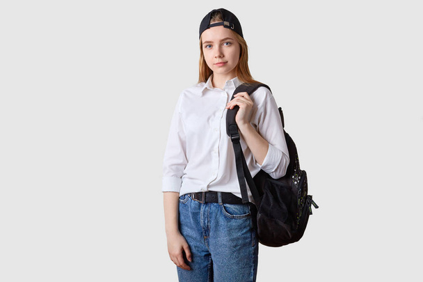 Image of beautiful student girl wearing fashionable jeans nad white shirt, walking to college from home, carrying backpack, looking directly at camera, posing against studio wall with copy space. - Foto, Imagem