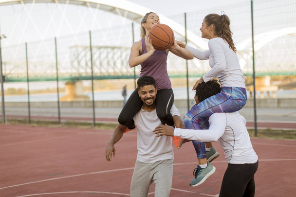 Jonge vrouwen die op de schouders van de mannen zitten en op een zonnige dag een basketbal houden op het buitenveld - Foto, afbeelding