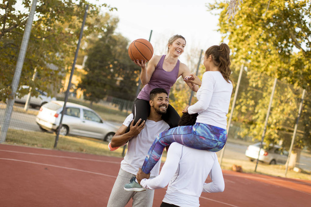Genç kadınlar erkeklerin omuzlarında oturuyor ve güneşli bir günde açık hava sahasında basketbol topunu tutuyorlar. - Fotoğraf, Görsel