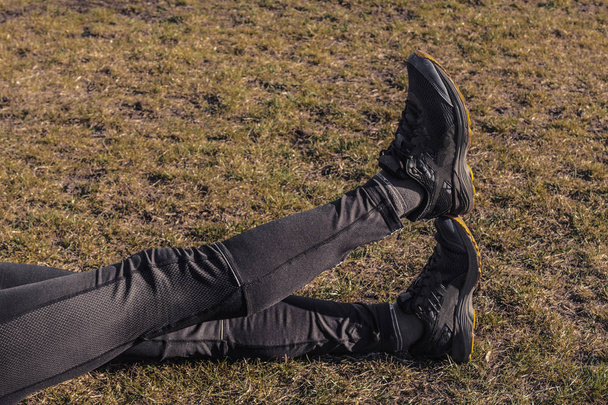 athlete's legs in leggings and sneakers, lying on the ground on the lawn close-up. crossed legs. training on the outdoors. warm-up, stretching - Foto, immagini