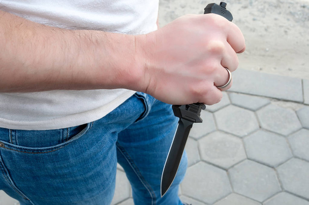 Ένας άντρας με μπλε τζιν και ένα λευκό μπλουζάκι με ένα μαύρο μαχαίρι τσέπης στο χέρι του. - Φωτογραφία, εικόνα