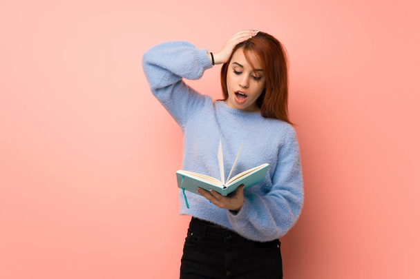 Young redhead woman over pink background surprised while enjoying reading a book - Φωτογραφία, εικόνα