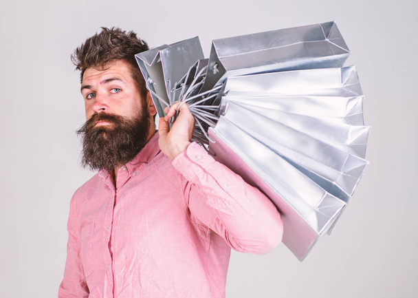 Shopping concept. Guy shopping on sales season, carries bunch of bags on shoulder. Man with beard and mustache holds shopping bags, grey background. Hipster on serious face shopping - Zdjęcie, obraz