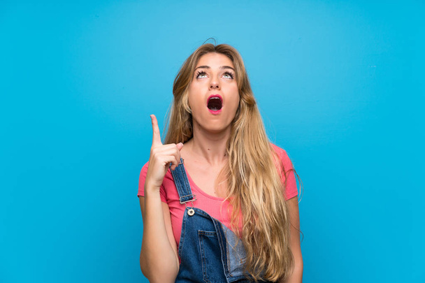Young blonde woman with overalls over isolated blue wall pointing up and surprised - Φωτογραφία, εικόνα