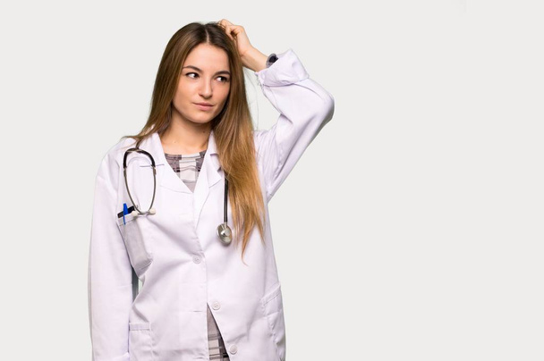 Young doctor woman having doubts while scratching head on isolated grey background - Fotoğraf, Görsel