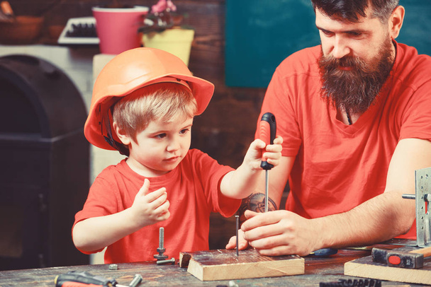 Father, parent with beard teaching little son to use tool screwdriver. Boy, child busy in protective helmet learning to use screwdriver with dad. Repair and workshop concept - Foto, Imagen