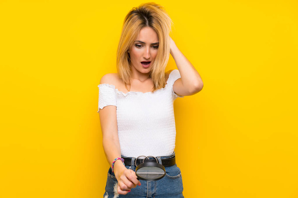 Young blonde woman over isolated yellow wall holding a magnifying glass - Photo, image