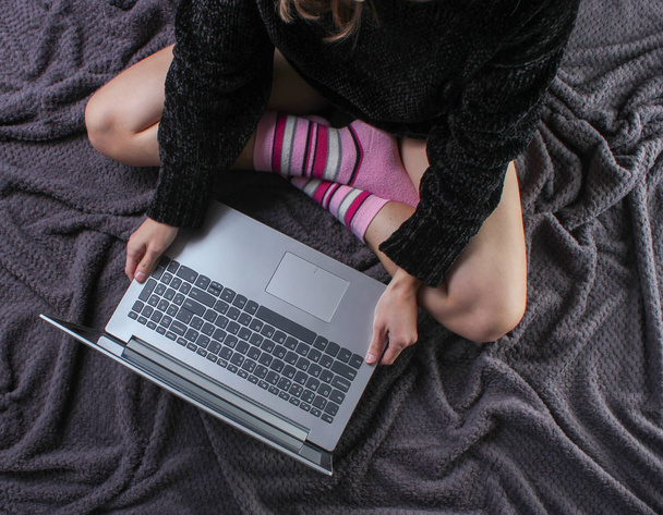 Damskie nogi siedzą na kocu i korzystają z laptopa. Zimowy poranek, przestrzeń robocza. Przytulne i ciepłe miejsce pracy, praca online. Widok z góry - Zdjęcie, obraz