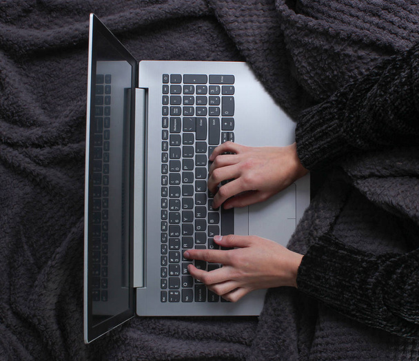 Le gambe femminili avvolte nella coperta si siedono e usano un computer portatile. Mattina invernale, spazio di lavoro. Luogo di lavoro accogliente e caldo, lavoro online. Vista dall'alto
 - Foto, immagini