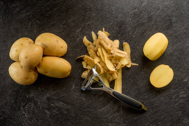 Peeler with peeled potatoes and their skins on textured black background. - Photo, Image