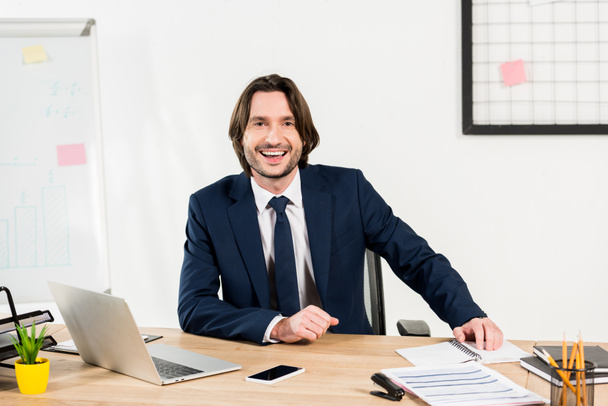 hombre alegre en ropa formal sonriendo cerca de la computadora portátil y el teléfono inteligente en la oficina
  - Foto, Imagen