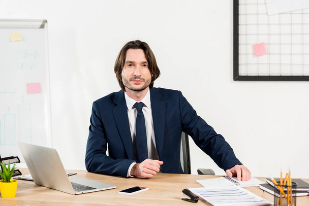 handsome man in formal wear sitting near laptop and smartphone in office  - Foto, Bild
